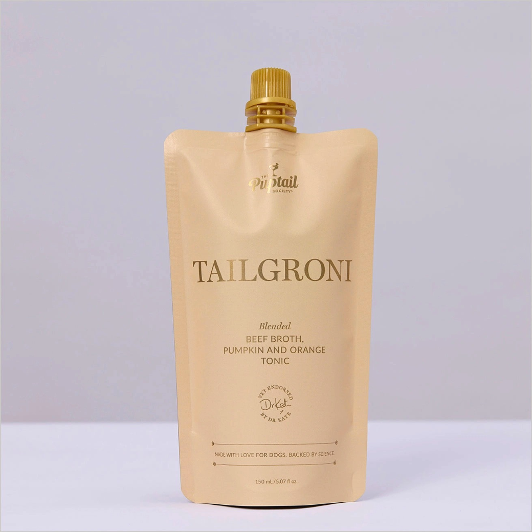 Coco & Pud - Tailgroni - Blended Beef Broth, Pumpkin and Orange Tonic Mocktail - Puptail Society. A 150 mL stand-up pouch of TAILGRONI dog tonic in a warm beige colour with a gold cap.