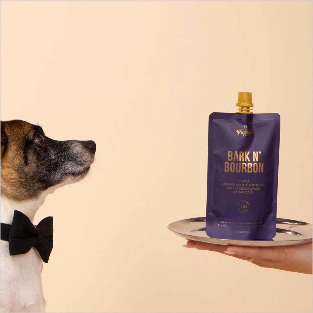 Coco & Pud - A Jack Russell dog wearing a black bow tie looks toward a purple pouch of Bark N' Bourbon dog tonic held on a silver tray - Puptail Society