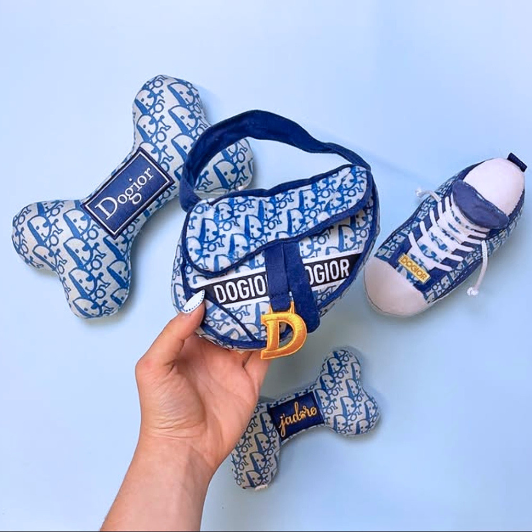 Coco & Pud - Dogior saddlebag plush dog toy in front of three matching plush toys shaped like a bone and a sneaker, all in a blue and white iconic French Dior Oblique pattern - Haute Diggity Dog