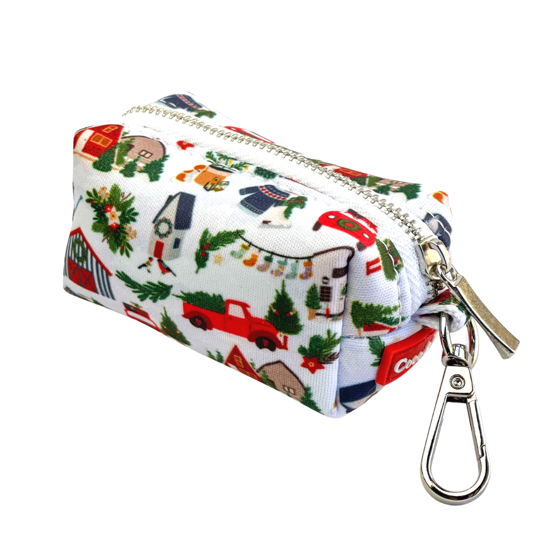 Coco & Pud - Home For Christmas, waste bag holder with a colourful Christmas-themed print featuring red trucks, green trees, andChristmas decorations. It has a silver metal zipper and a silver carabiner clip attached.