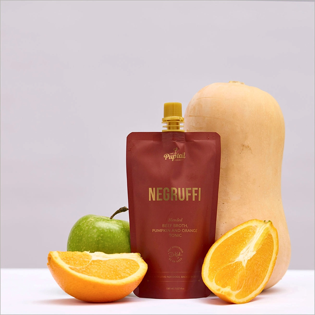 Coco & Pud - A pouch of Negruffi blended beef broth, pumpkin and orange tonic for dogs stands on a white surface with a whole butternut squash, a green apple, and two orange slices arranged around it. Puptail Society