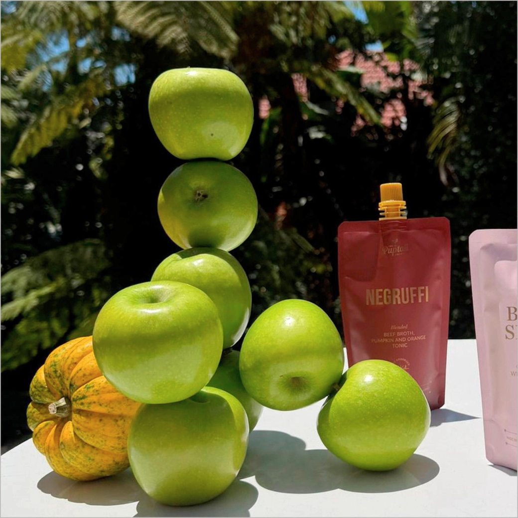 Coco & Pud - Negruffi blended beef broth, pumpkin and orange tonic for dogs. A stack of bright green apples and a small pumpkin sit on a white table beside two pouches, one dark red labelled NEGRUFFI and one pale pink partially visible - Puptail Society