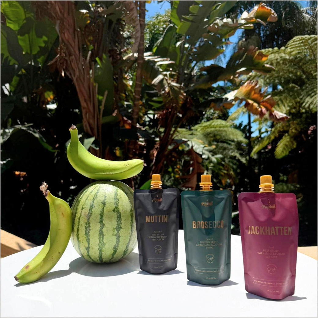 Coco & Pud - 3 pouches of Puptail Society Muttini, Brosecco, Jackhatten tonics for dogs arranged on a white table outdoors with bananas and a watermelon