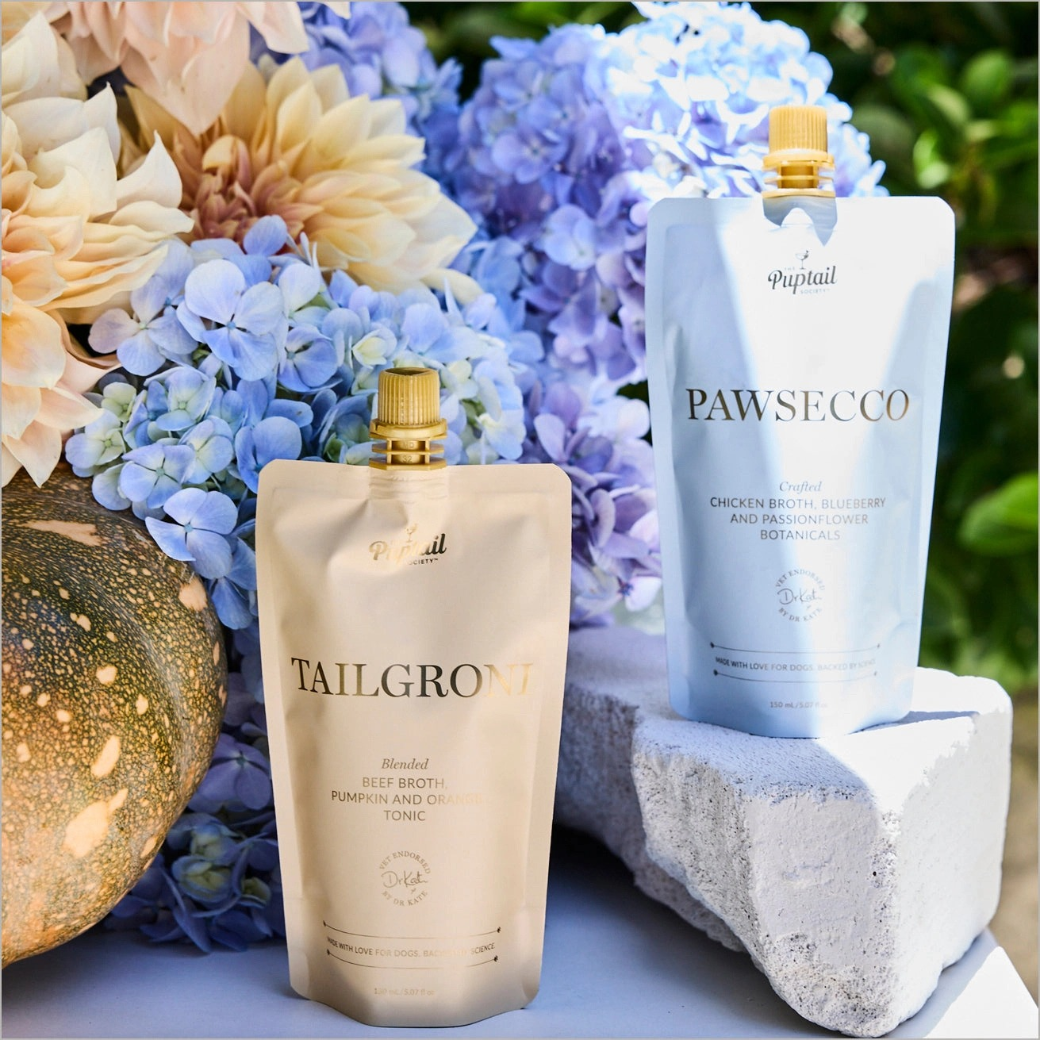 Coco & Pud - Tailgroni & Pawsecco Puptail pet supplement pouches, Tailgroni in cream and Pawsecco in light blue, styled with hydrangeas and a pumpkin in an outdoor garden setting -  Puptail Society