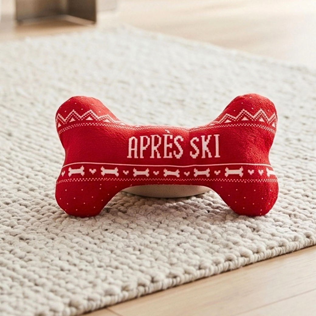 Coco & Pud - Apres Ski red bone-shaped dog toy with white knit-style patterns and the text 'APRÈS SKI' on the front, placed on a cream textured rug over a light wood floor - Haute Dggity Dog