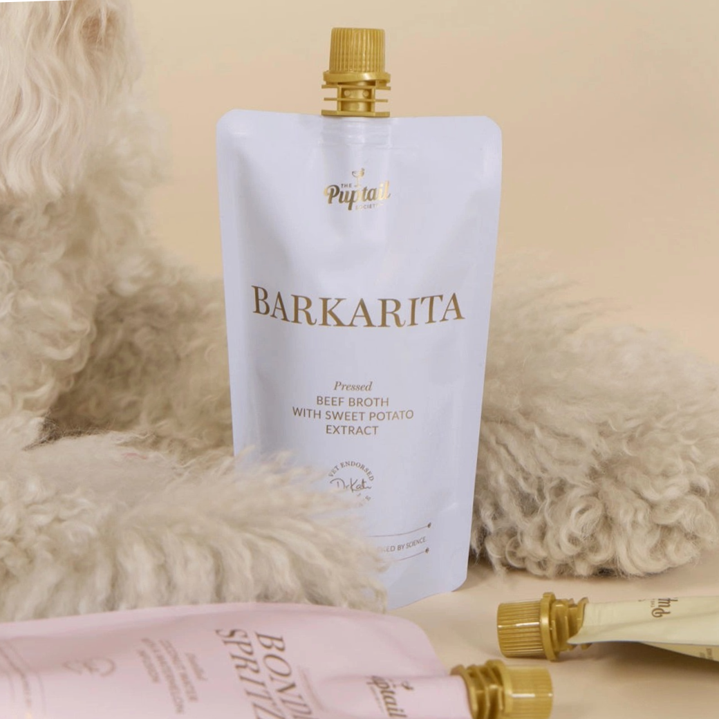 Coco & Pud - A white stand-up pouch labelled Barkarita Beef Broth with Sweet Potato Extract stands upright on a beige surface, surrounded by soft white faux fur - Puptail Society