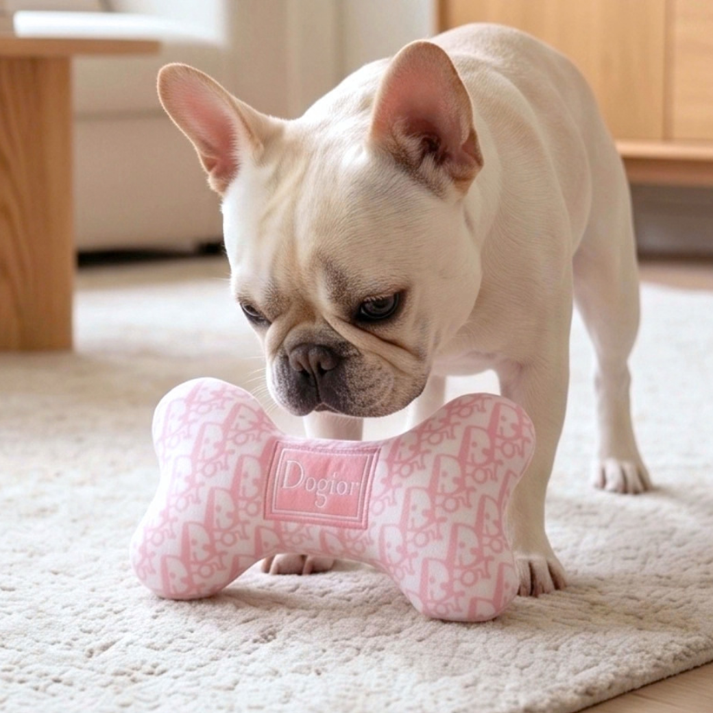 A French Bulldog stands looking down at a pink and white bone-shaped dog toy with a repeating pattern and a central label that reads 'Dogior' - Haute Diggity Dog