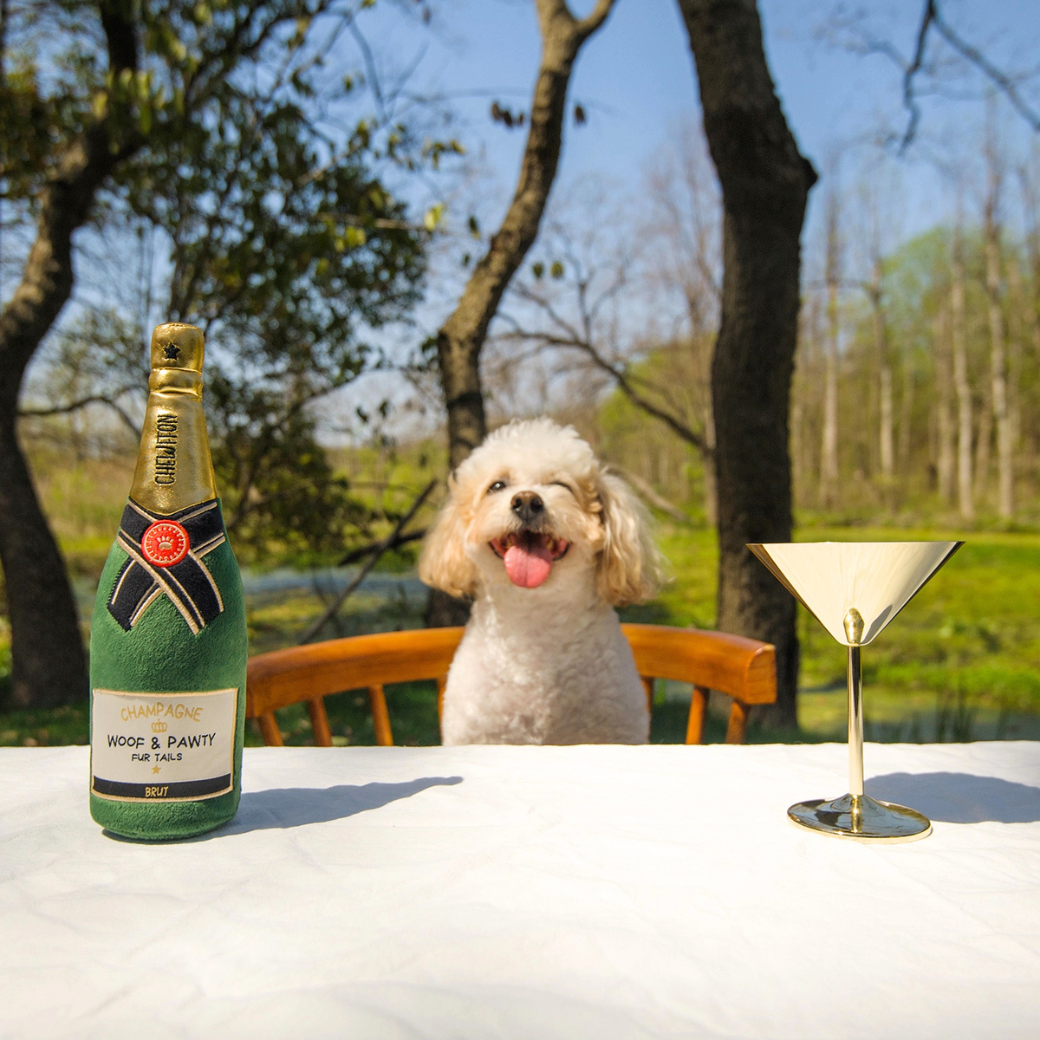 Coco & Pud - Woof & Pawty Champagne dog toy with a small white dog sits on a wooden chair behind a table outdoors, with a green bottle of Wood & Pawty and a gold martini glass - Chewffon