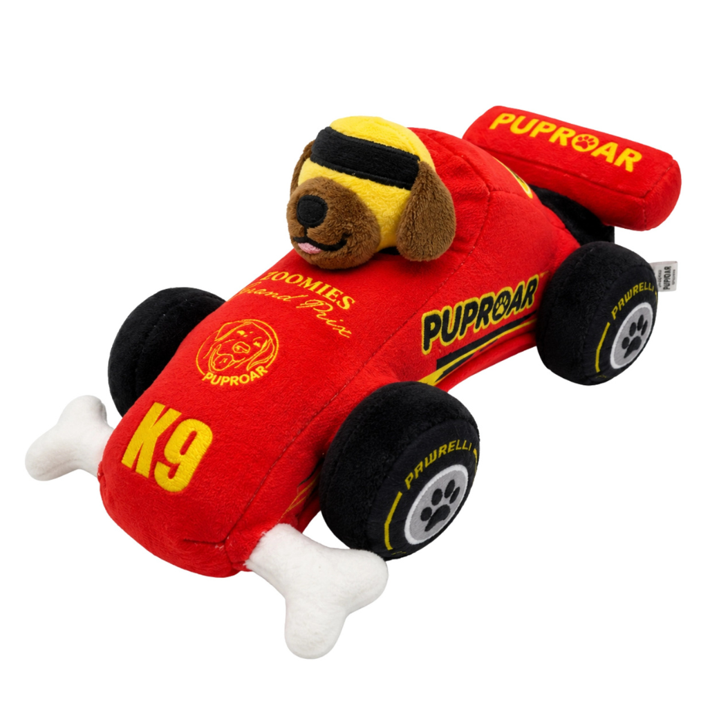 Coco & Pud - Puppy Prix Speeder Racecar plush dog toy. A plush racing dog toy shaped like a red race car with a brown dog head wearing yellow goggles, black wheels, and white bone-shaped front feet. Visible text includes 'PUPROAR', 'K9' - Puproar