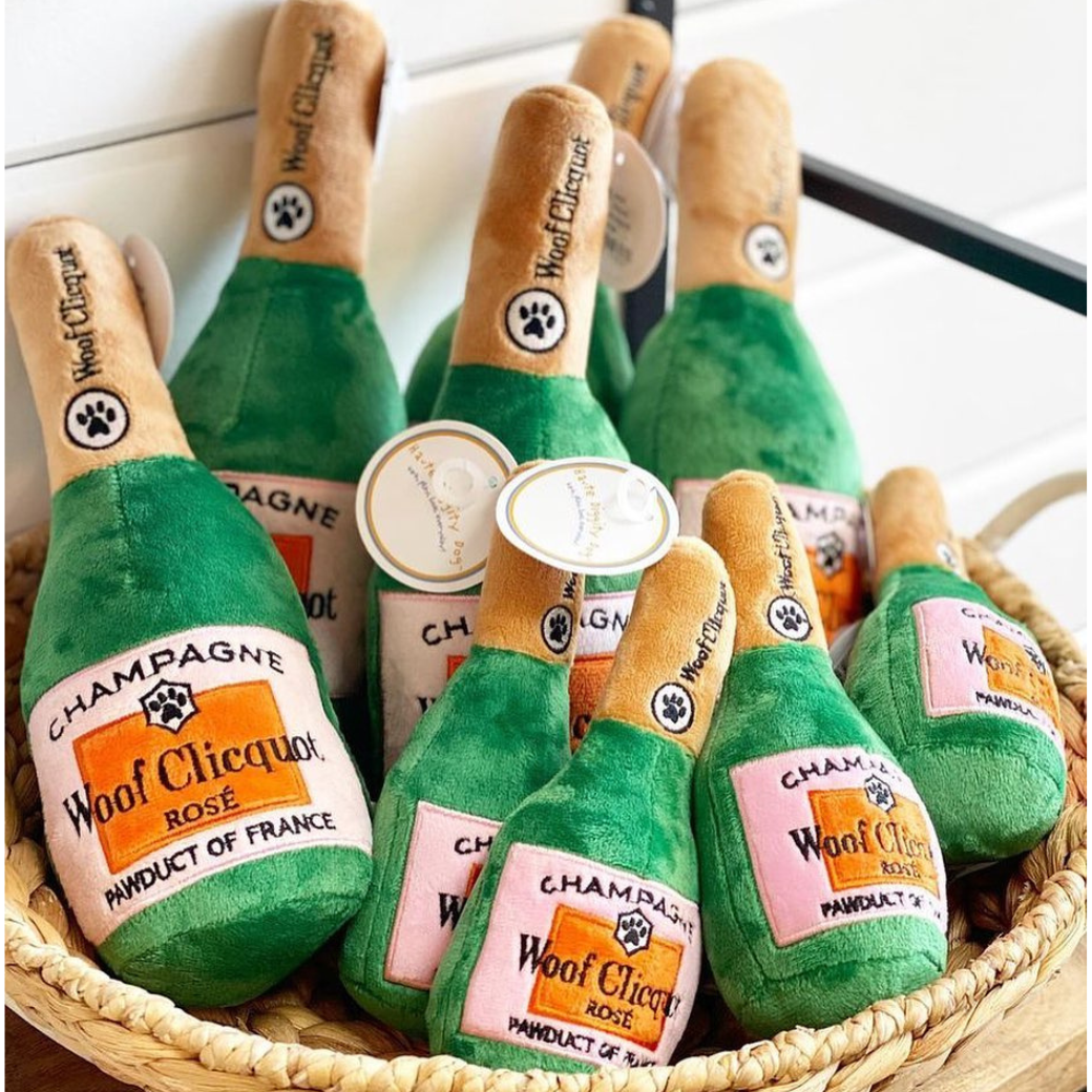 Coco & Pud - A basket of plush dog toys shaped like champagne bottles, labelled 'Champagne' and 'Woof Clicquot Rose,' small and large sizes.